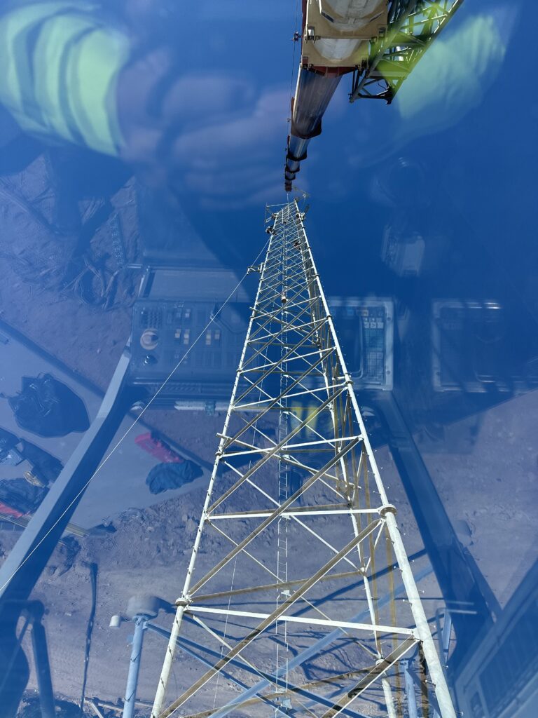 Crane boom lifting a tall metal tower structure against clear blue sky